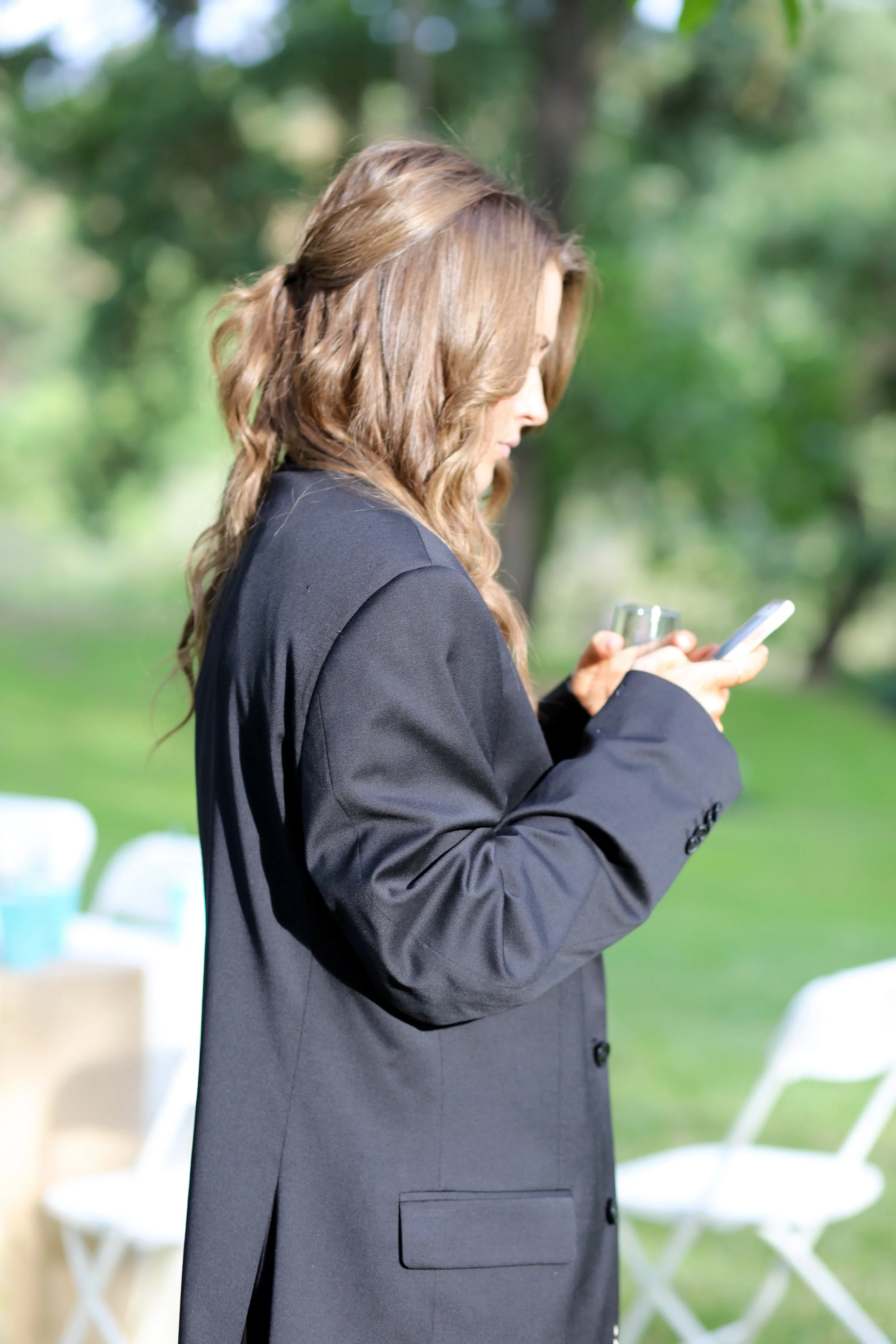 A woman with wavy brown hair, wearing a dark gray blazer, and holding a smartphone, stands outdoors with blurred green trees and white chairs in the background.