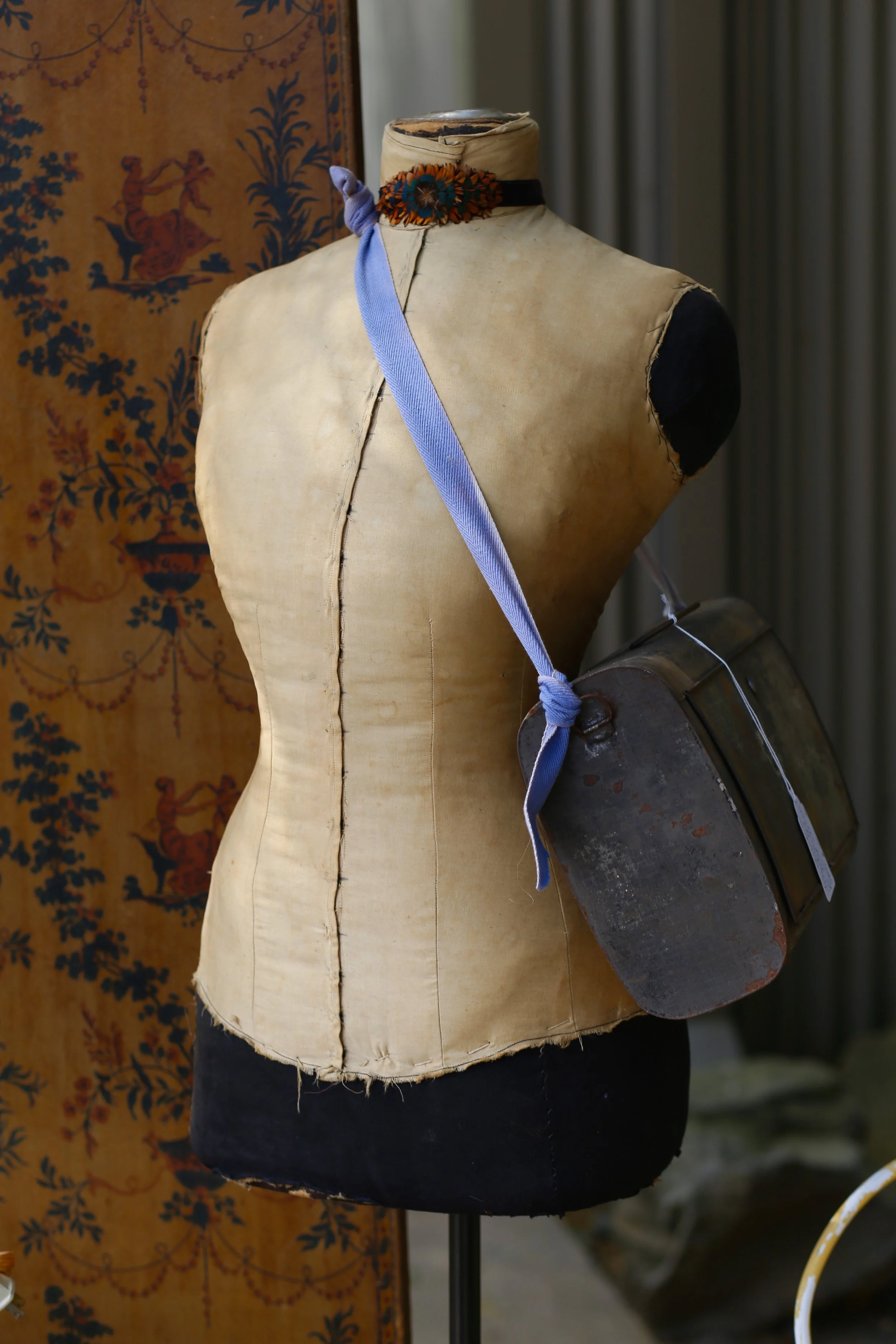 An antique dress form mannequin with a beige fabric cover, wearing a decorative choker made of fabric flowers, and a purple ribbon tied in a bow. The mannequin holds a rusty, vintage metal lunchbox or container. A tapestry with a floral pattern is vi