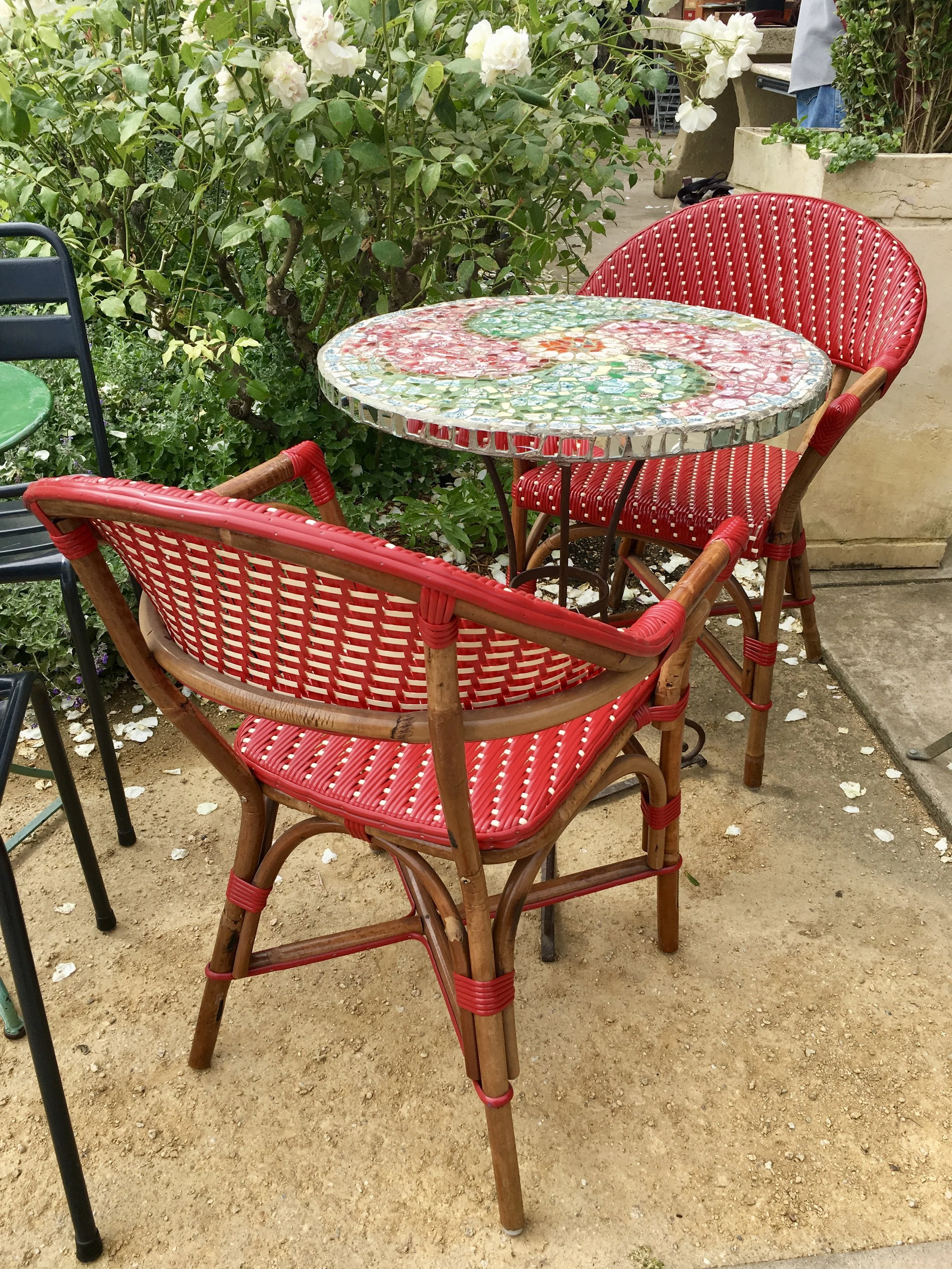 A round, mosaic-topped table with a red and green design, surrounded by red wicker chairs with wooden frames, set outdoors on a dirt surface near green bushes and potted plants.