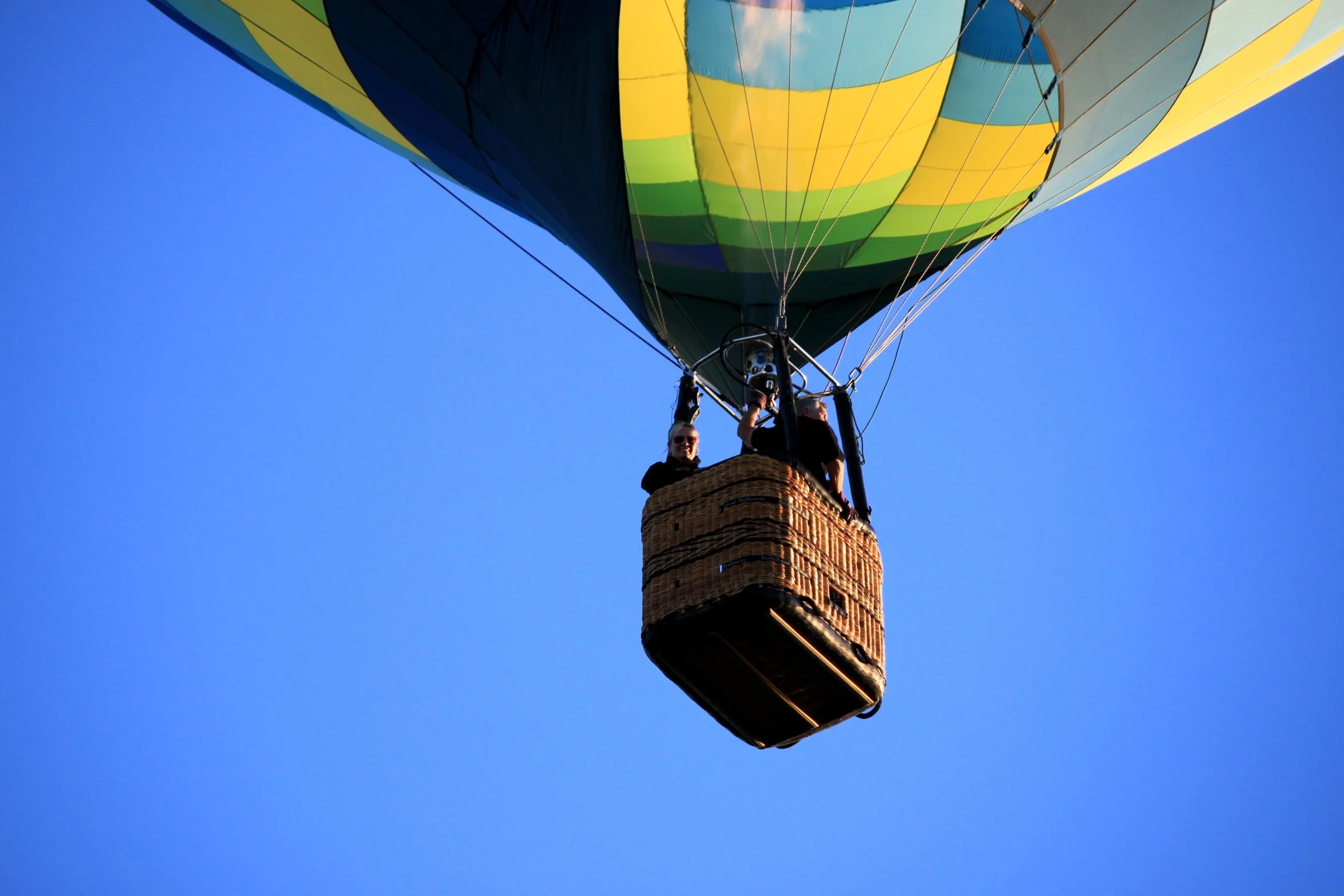 A hot air balloon with a multicolored pattern of blue, yellow, green, and white, flying in a clear blue sky. There are two passengers in the basket, one woman smiling and waving, and one man looking inside the basket in Yountville CA