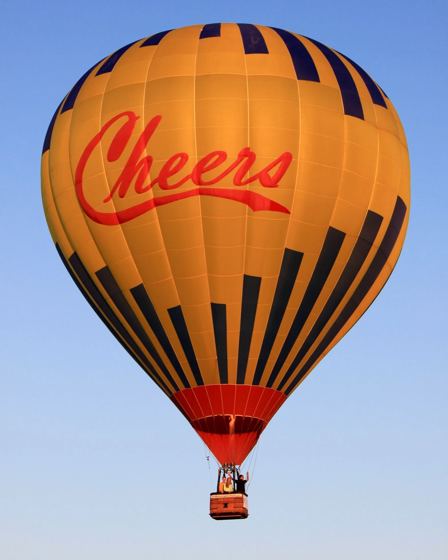Yellow and black hot air balloon with the word "Cheer" written in red on the side, floating in clear blue sky with two people visible in the basket.