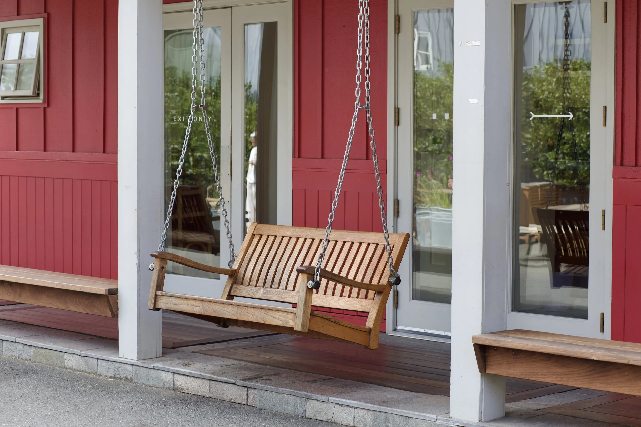 A wooden porch swing hanging from chains outside a red building with white trim and glass doors.