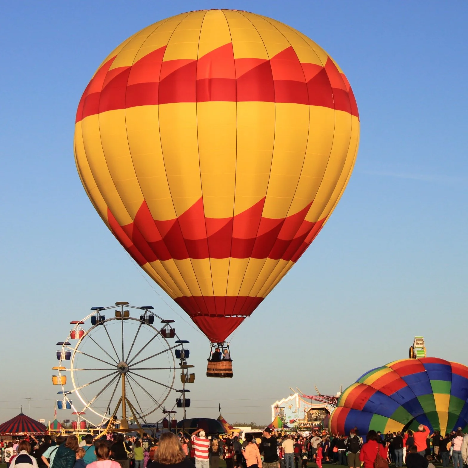 A hot air balloon with red and yellow patterns floating above a crowd at a fair, with a Ferris wheel in the background and a clear blue sky.