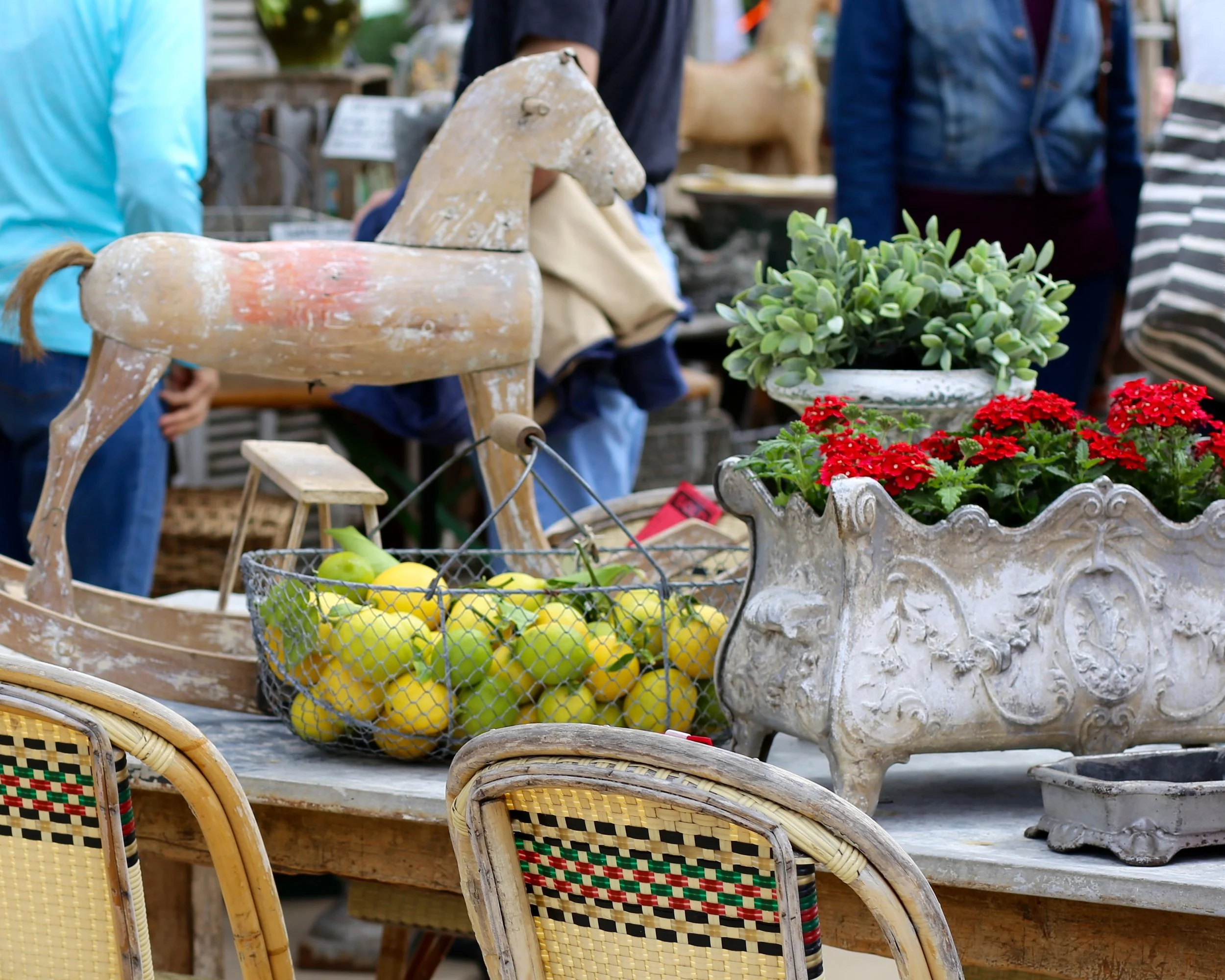 A market scene with baskets of green and yellow lemons, potted plants, and vintage decorative items. Several people are in the background browsing the items.