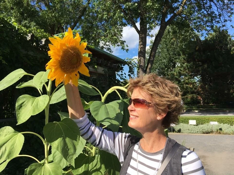 A woman with short curly hair and sunglasses smiling and touching a large sunflower in a garden on a sunny day.