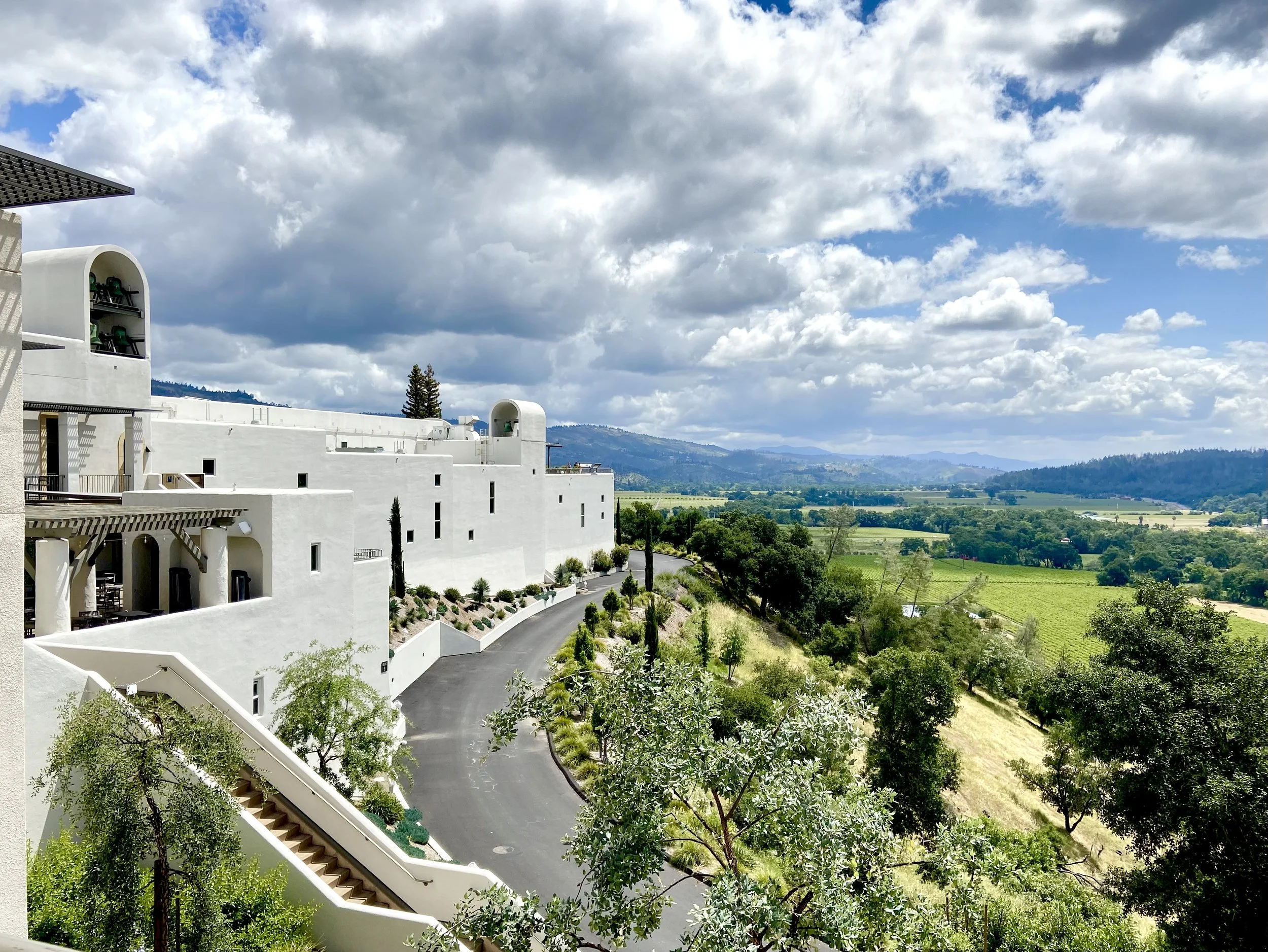A white building with modern architectural style, featuring multiple levels and terraces, is situated on a hill amid lush greenery. The background shows a vast landscape of fields, trees, and distant mountains under a partly cloudy sky with shades of