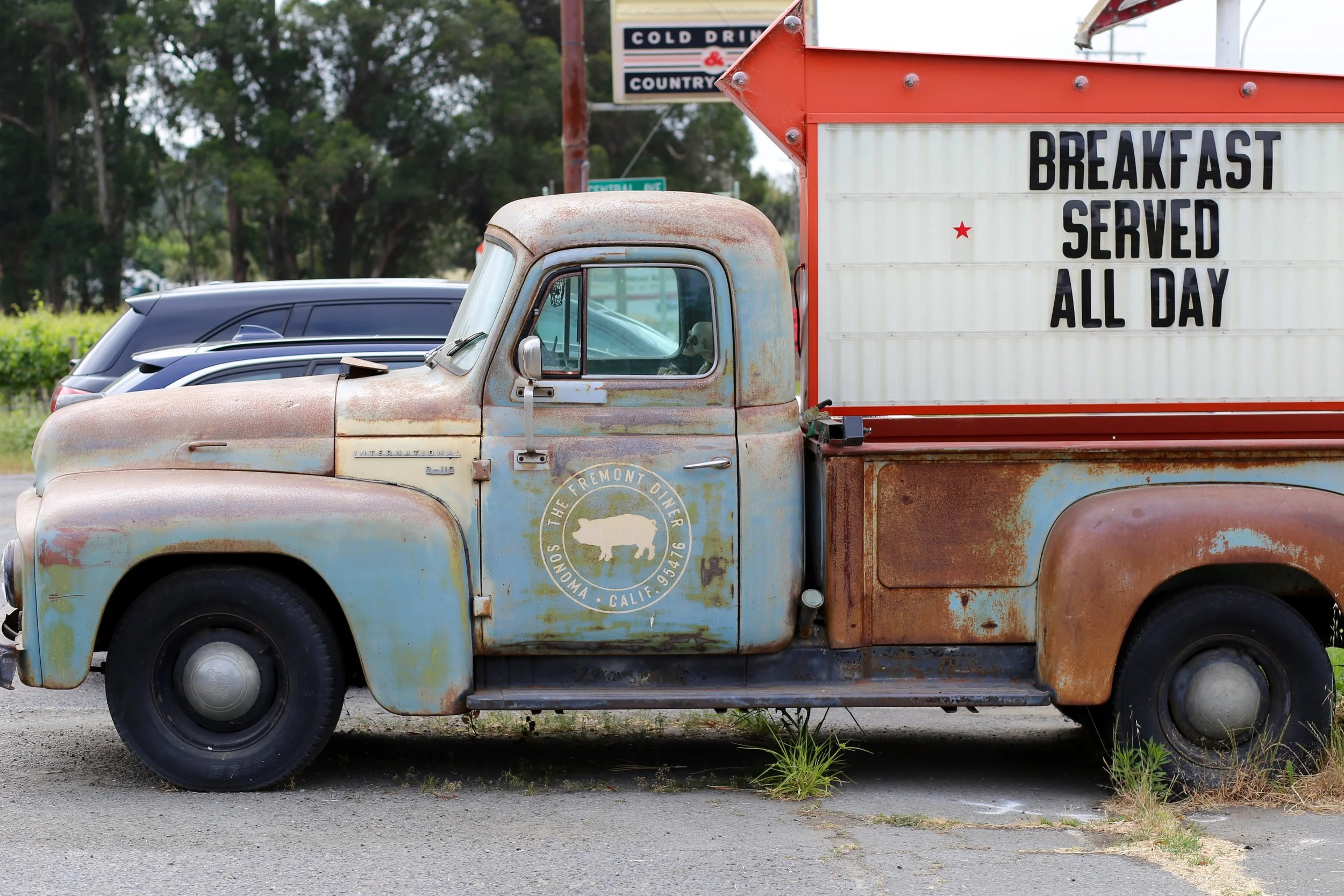 An old, rusty pickup truck parked in a lot, with a sign on the side advertising breakfast served all day and a skeleton inside the cab.