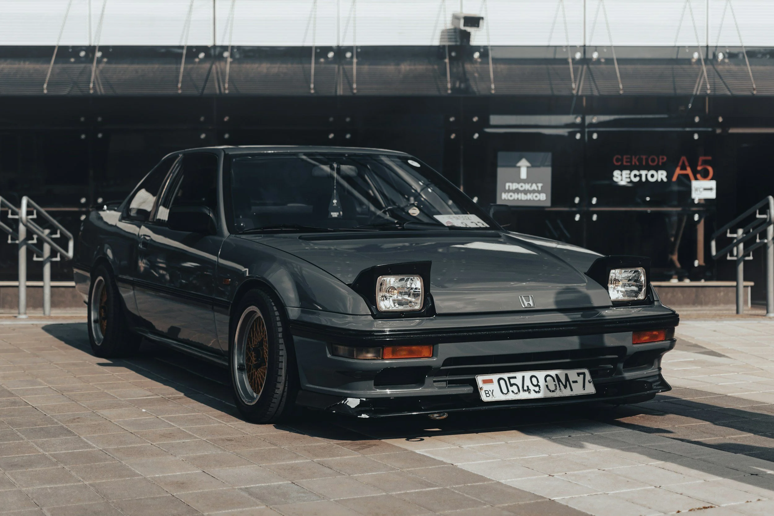 A black vintage Honda sports car parked outside on a tiled pavement with signs in the background indicating a parking area, sector A5, and directions in Cyrillic script.