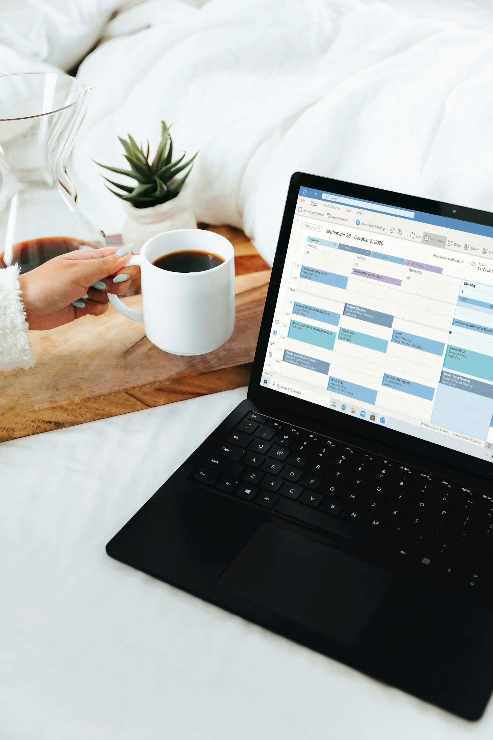 A laptop on a white surface displaying a digital calendar with scheduled appointments. A hand holds a white coffee mug filled with black coffee. In the background, there is a glass carafe with coffee and a small potted succulent plant. The scene is cozy and organized.
