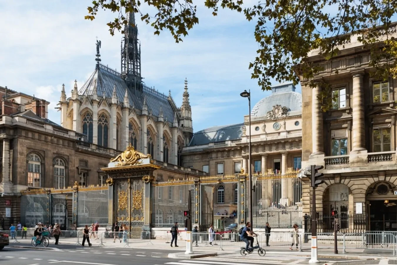 Street view of a historic building with ornate architecture, including a tall spire and decorative gold accents, surrounded by a gold fence and busy pedestrians and cyclists.