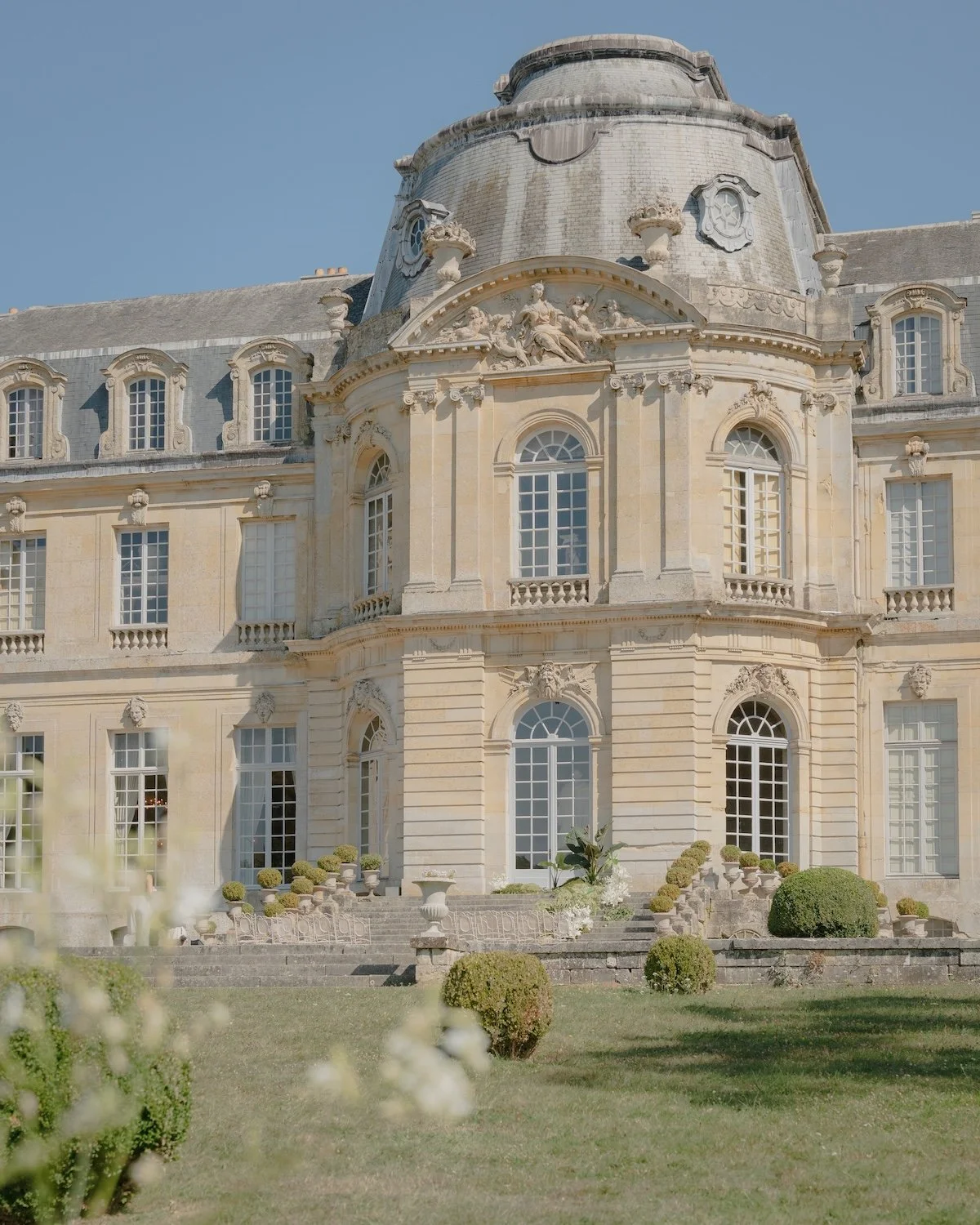 A grand, historic European-style mansion with large windows, ornate architectural details, and a garden with trimmed bushes in front.