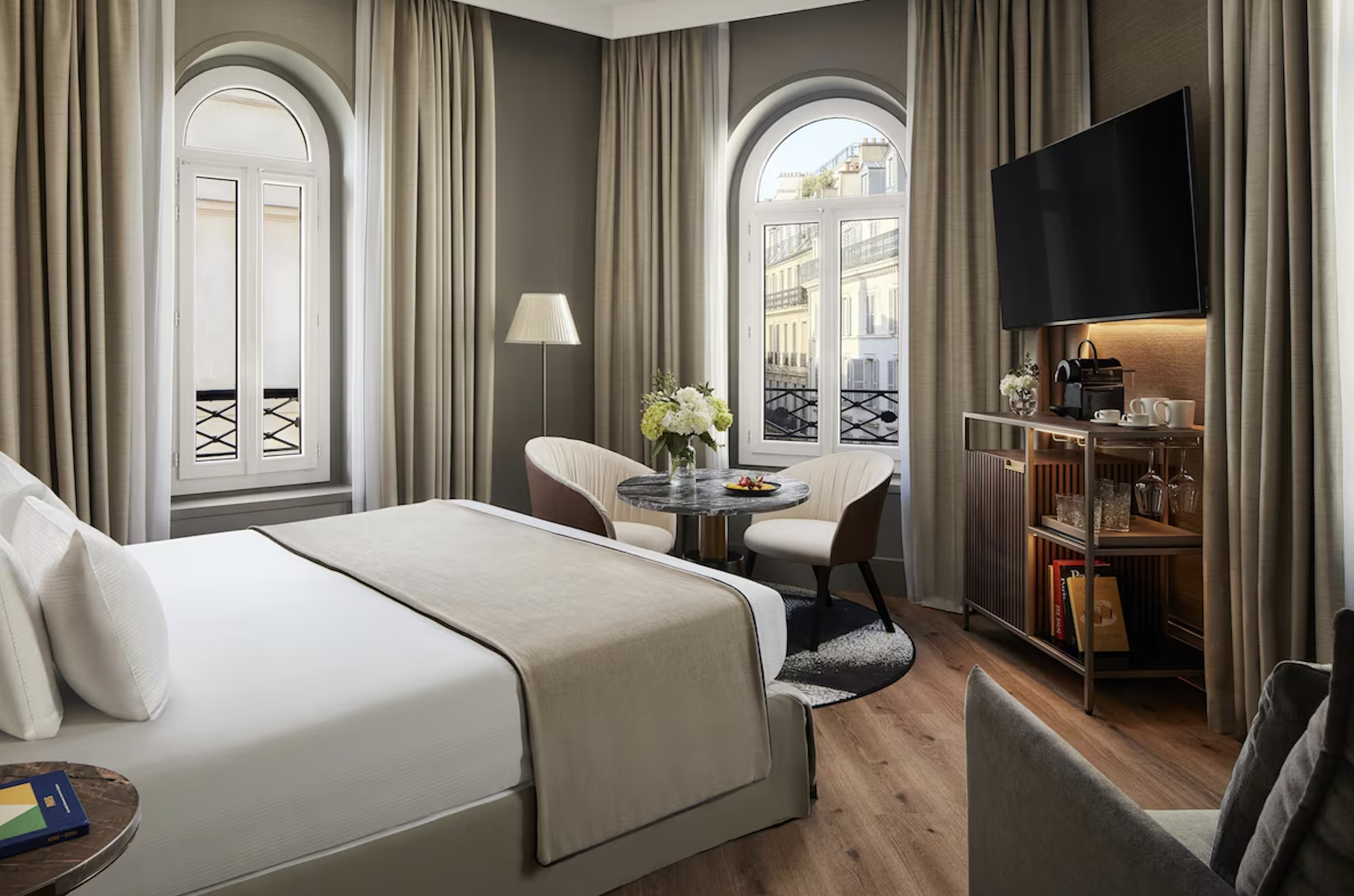 A hotel room with large arched windows, cream curtains, a white bed, a nightstand, a round table with two chairs, a lamp, a bouquet of white flowers, a flat-screen TV, a coffee station, and a wooden bookshelf with books.