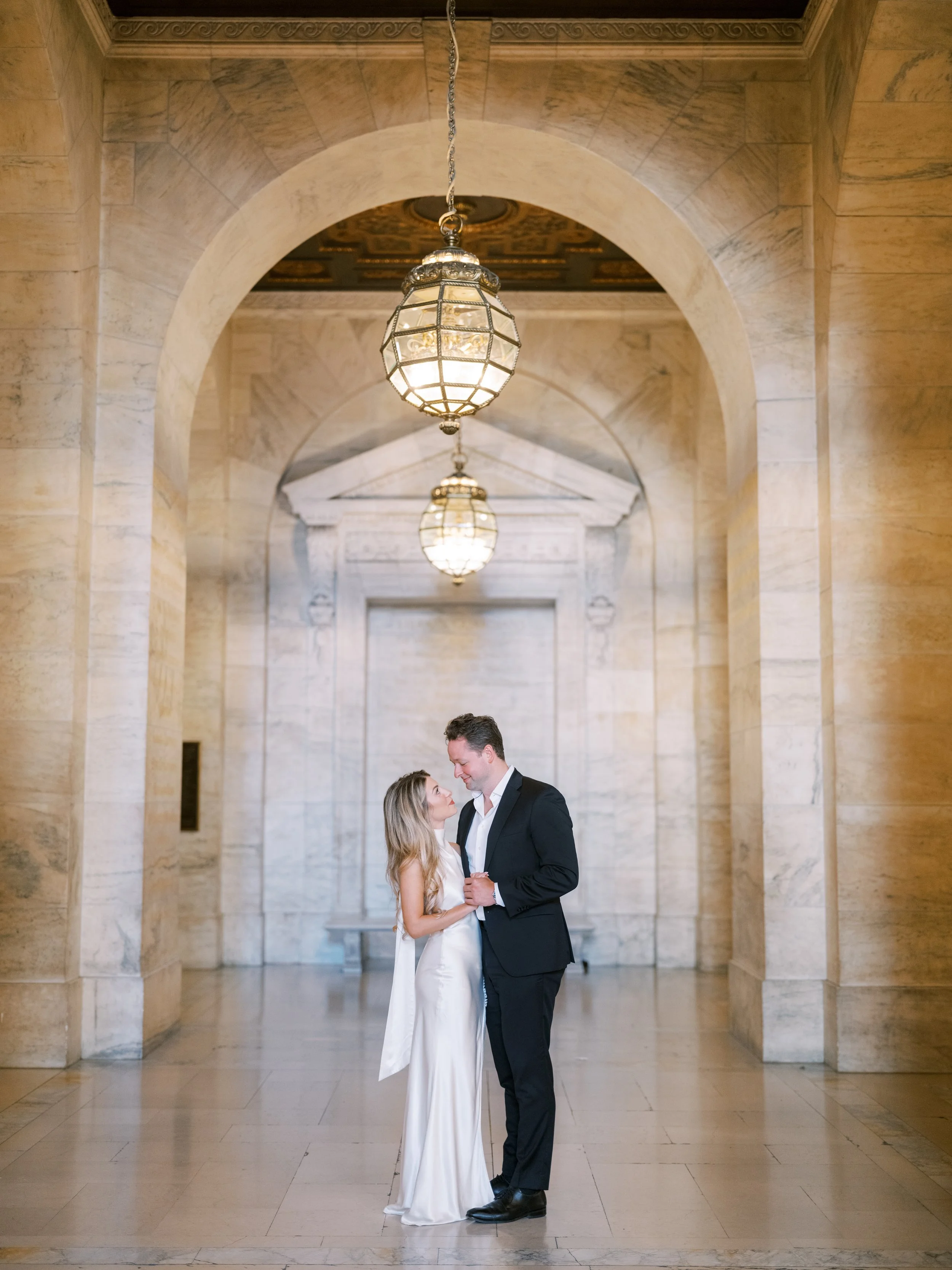 A romantic moment between a man in a suit and a woman in a white dress, standing close together under a chandelier in an elegant marble hallway. New York Public Library. NYPL. Samantha Daniell & Michael Moore. 