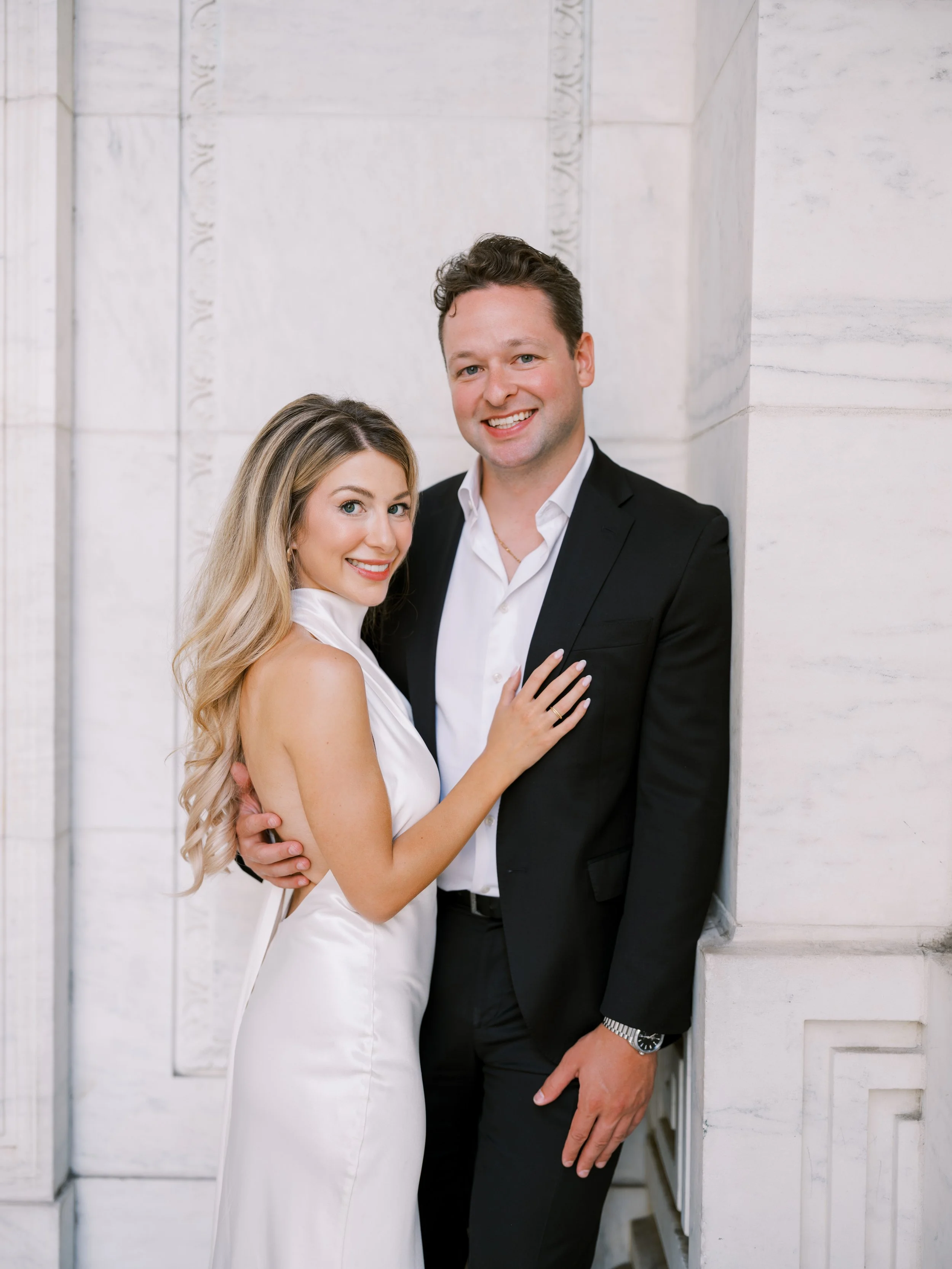 A smiling man in a black suit with a white shirt standing next to a smiling woman in a white dress, both standing against a white stone wall.