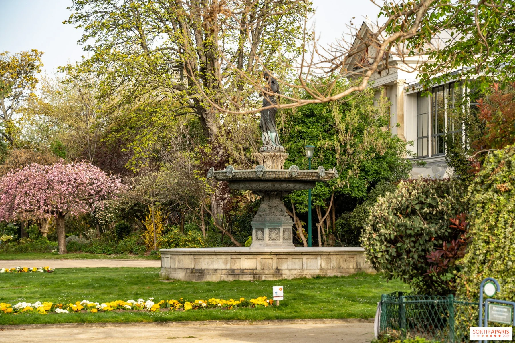 A decorative fountain with a statue on top, surrounded by colorful trees and shrubs in a garden. There are flowers and a sign in front of the fountain, and a bench to the right.