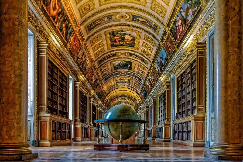 A grand, historic library hall with ornate gold-accented ceiling paintings and tall wooden bookshelves lining each side. A large globe sculpture is centered in the room.