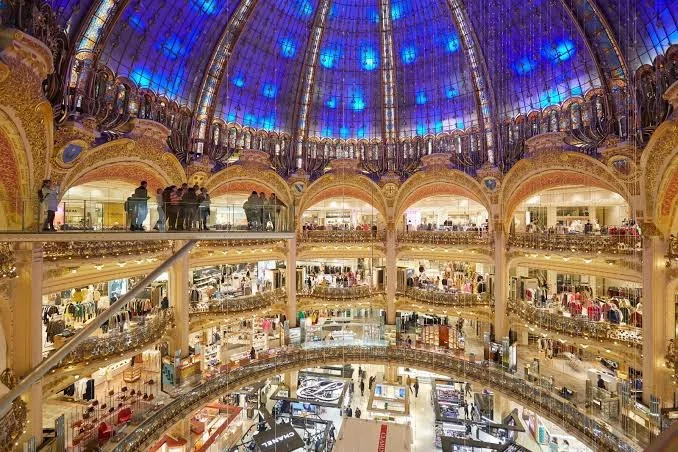 Interior of a luxury shopping mall with a large blue stained glass ceiling and multiple floors filled with stores and shoppers.