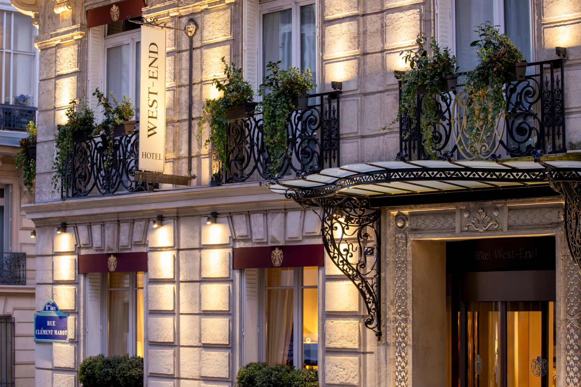 Exterior of the Hotel West-End with ornate black wrought iron balconies, several potted plants, and a vertical sign for the hotel. The building has beige stone walls illuminated by exterior lighting, with multiple windows and decorative architectural details.