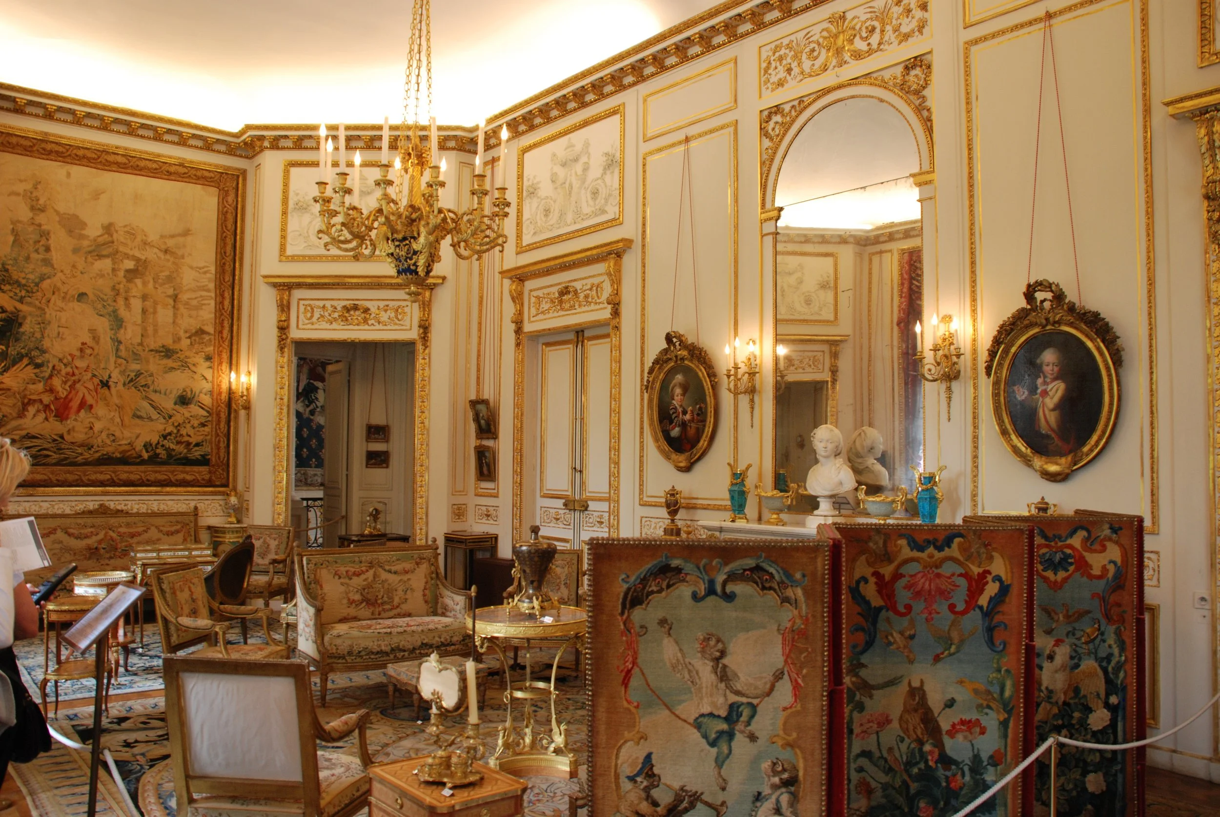 Luxurious room decorated with gold accents, ornate moldings, paintings, a chandelier, and antique furniture.