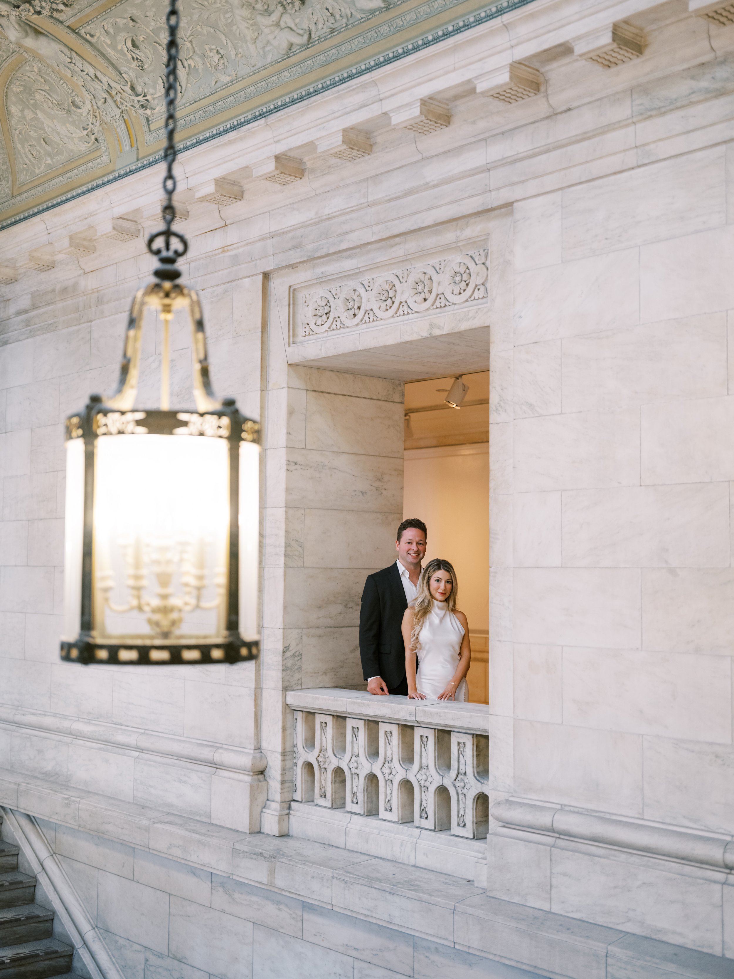 A man and a woman in formal attire standing on a marble balcony in a building with ornate architectural details. The man is wearing a suit, and the woman is wearing a white dress. New York Public Library. NYPL. Samantha Daniell & Michael Moore. 