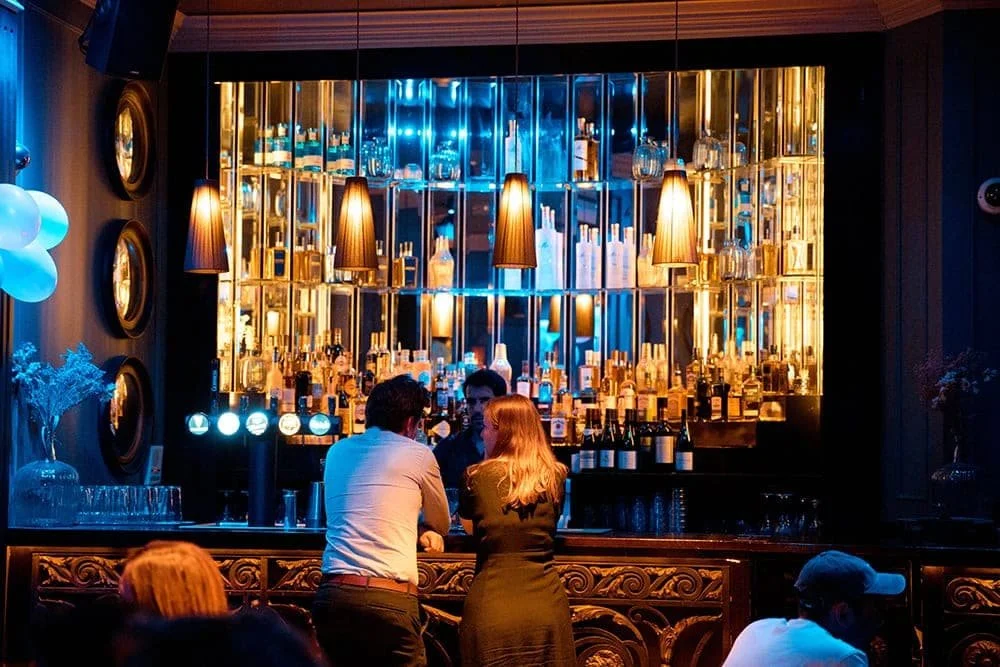 A bar with a backlit display of various bottles of alcohol, three pendant lights hanging above, and patrons engaging in conversation at the counter.
