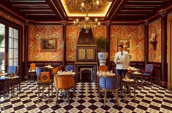 Elegant restaurant dining room with checkered black and white floor, ornate wallpaper, and antique furniture. A waiter is serving drinks, and the room has large windows, a chandelier, and decorative wall art.