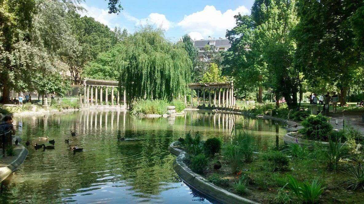 A park with a pond surrounded by greenery and trees, with some people walking and a few ducks swimming in the water.