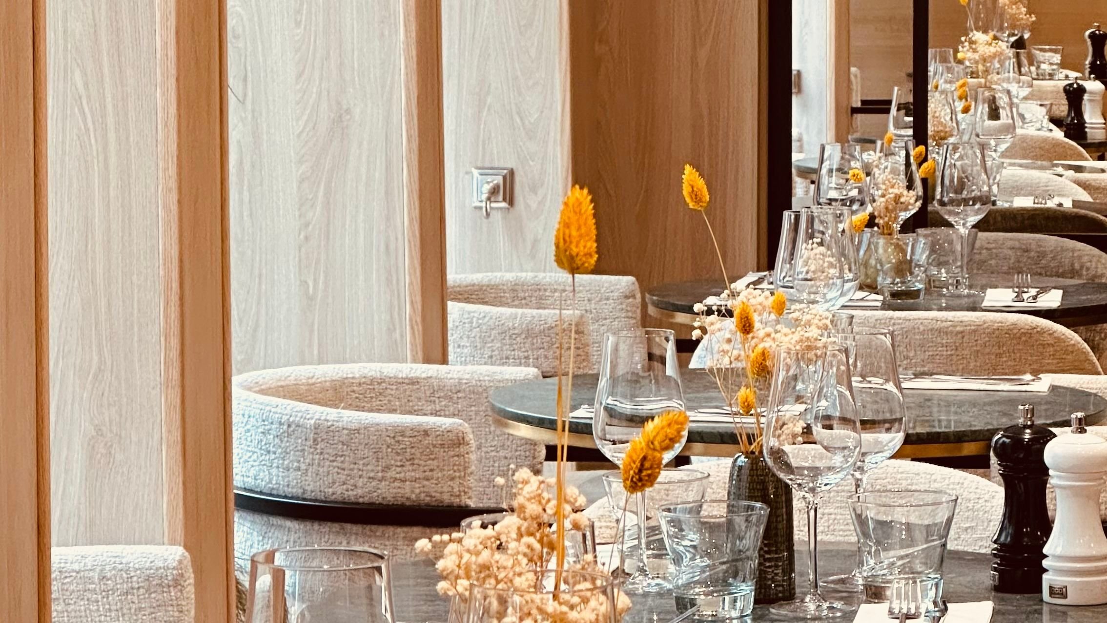 An elegant restaurant with round marble tables set with glassware, bottles, and floral centerpieces. Light-colored upholstered chairs surround the tables, and the walls are made of wood panels.