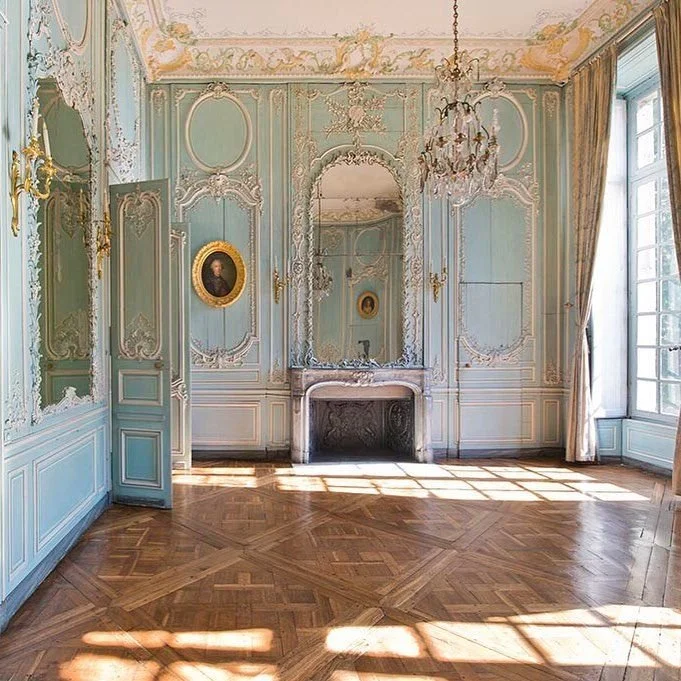 Elegant room with blue and white walls, intricate gold moldings, a large mirror above a marble fireplace, a chandelier hanging from the ceiling, and large windows with curtains letting in sunlight.