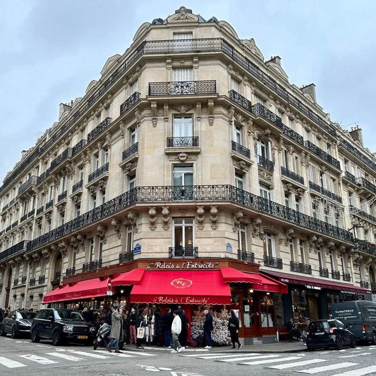 A corner building in Paris with intricate architectural details, featuring a ground-floor restaurant with red awnings and a crowd of people outside, and multiple upper floors with balconies and decorative elements.