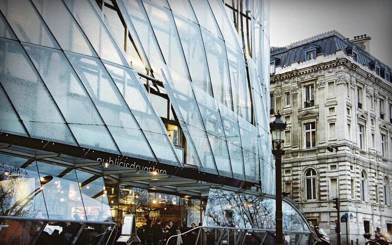 Modern glass building adjacent to a historic stone building in an urban street scene with street lamps and pedestrians.
