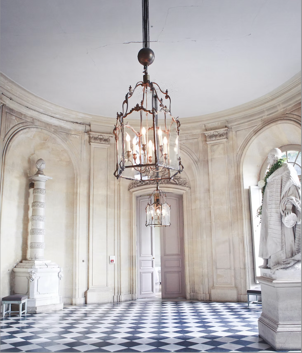 Elegant room with a checkered black and white floor, ornate chandeliers, sculpted walls, classical statues, and large arched windows.