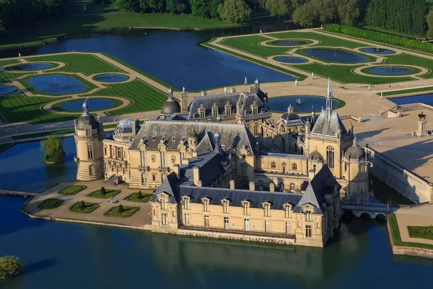 A large historic castle surrounded by water and a meticulously landscaped garden with circular and rectangular patterned pools and lawns, set in a lush green area.