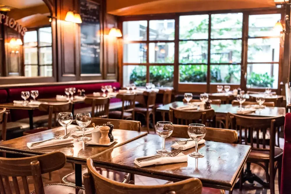 Empty restaurant dining area with wooden tables, chairs, and large windows. Glassware, napkins, and salt and pepper shakers are set on tables, with soft lighting and greenery outside.