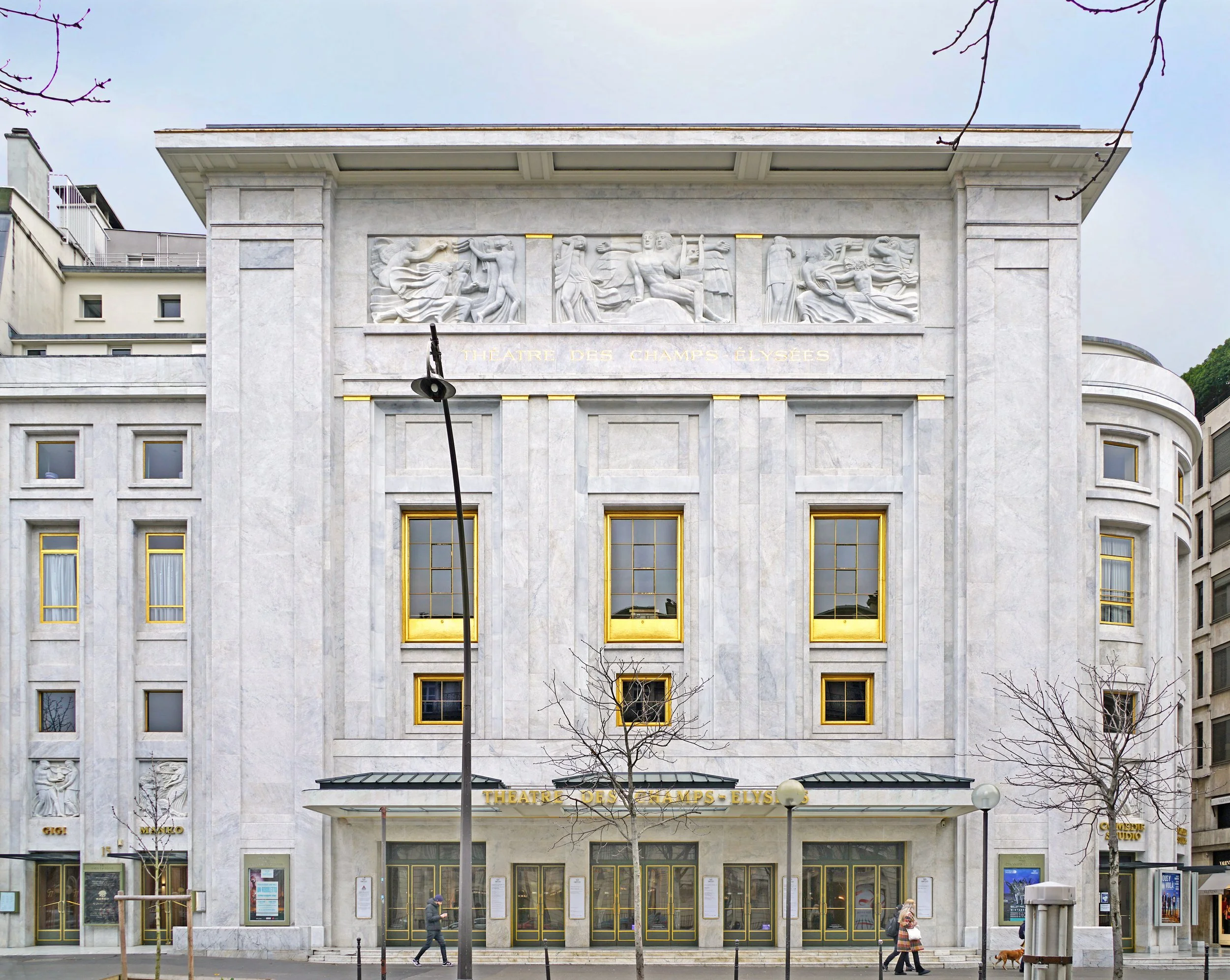 Front of the Théâtre des Champs-Élysées building with sculptures and gold accents, showing windows, trees, and people walking by.