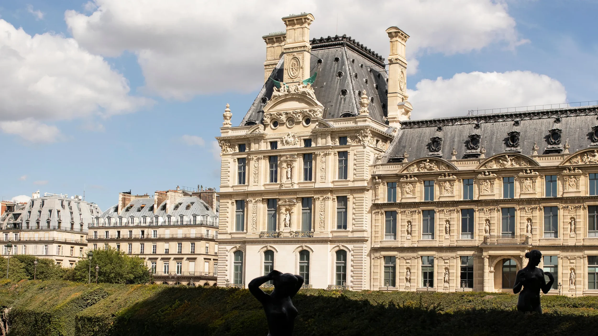 View of a historic Parisian building with ornate architecture, a steep mansard roof, and sculptures, with decorative statues and a chimneystack, under a partly cloudy sky. In the foreground, there are silhouetted statues in a garden and trees.