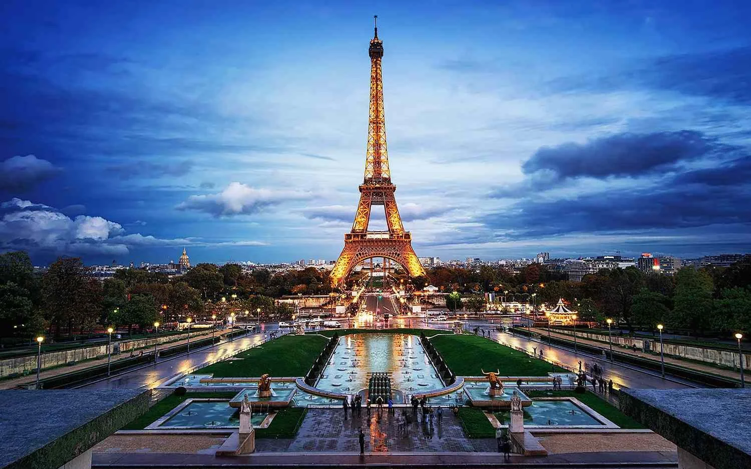 The Eiffel Tower illuminated at dusk in Paris, France, with a fountain and garden in the foreground and cityscape in the background.