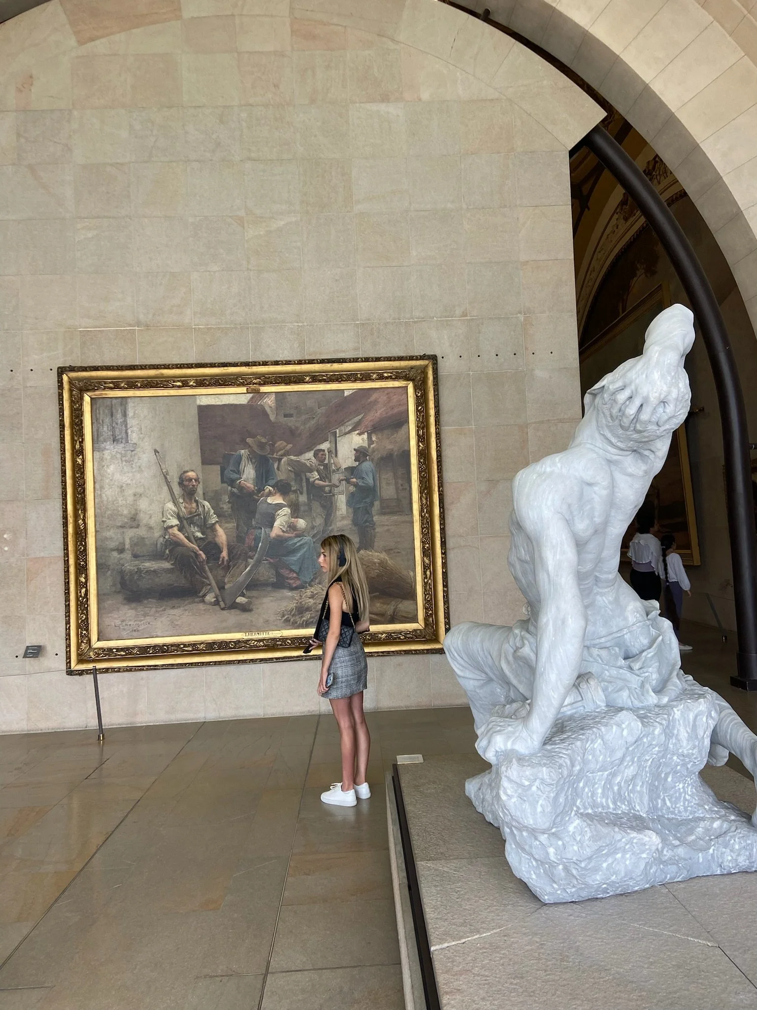 A woman with blonde hair and wearing a gray dress and white sneakers is standing in an art museum, looking at a large framed painting on the wall. The painting depicts a rustic scene with several men, some sitting and some standing, exchanging goods or ideas. To the right of the woman, there is a white marble sculpture of a muscular figure with a hand on its head, on a marble pedestal.