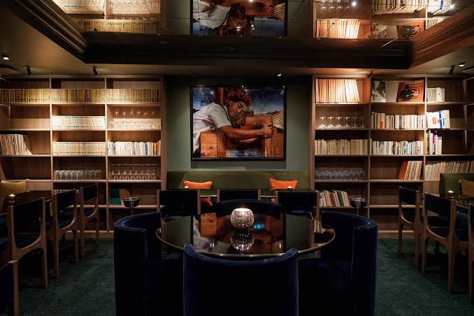 A cozy room with a round table surrounded by navy blue chairs, bookshelves filled with books along the walls, and a large framed photograph of a person resting on a wooden surface hanging on the wall.