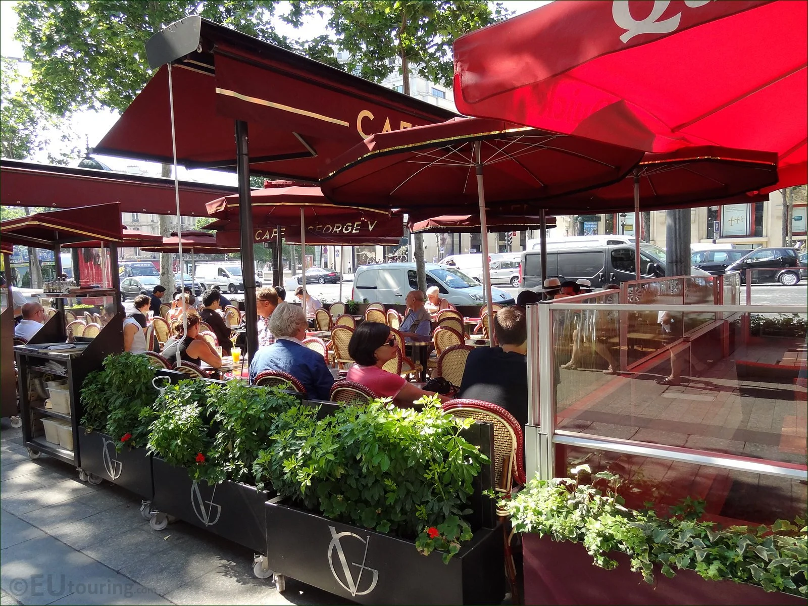 Outdoor cafe with red umbrellas and patio seating filled with people, surrounded by potted plants during daytime.