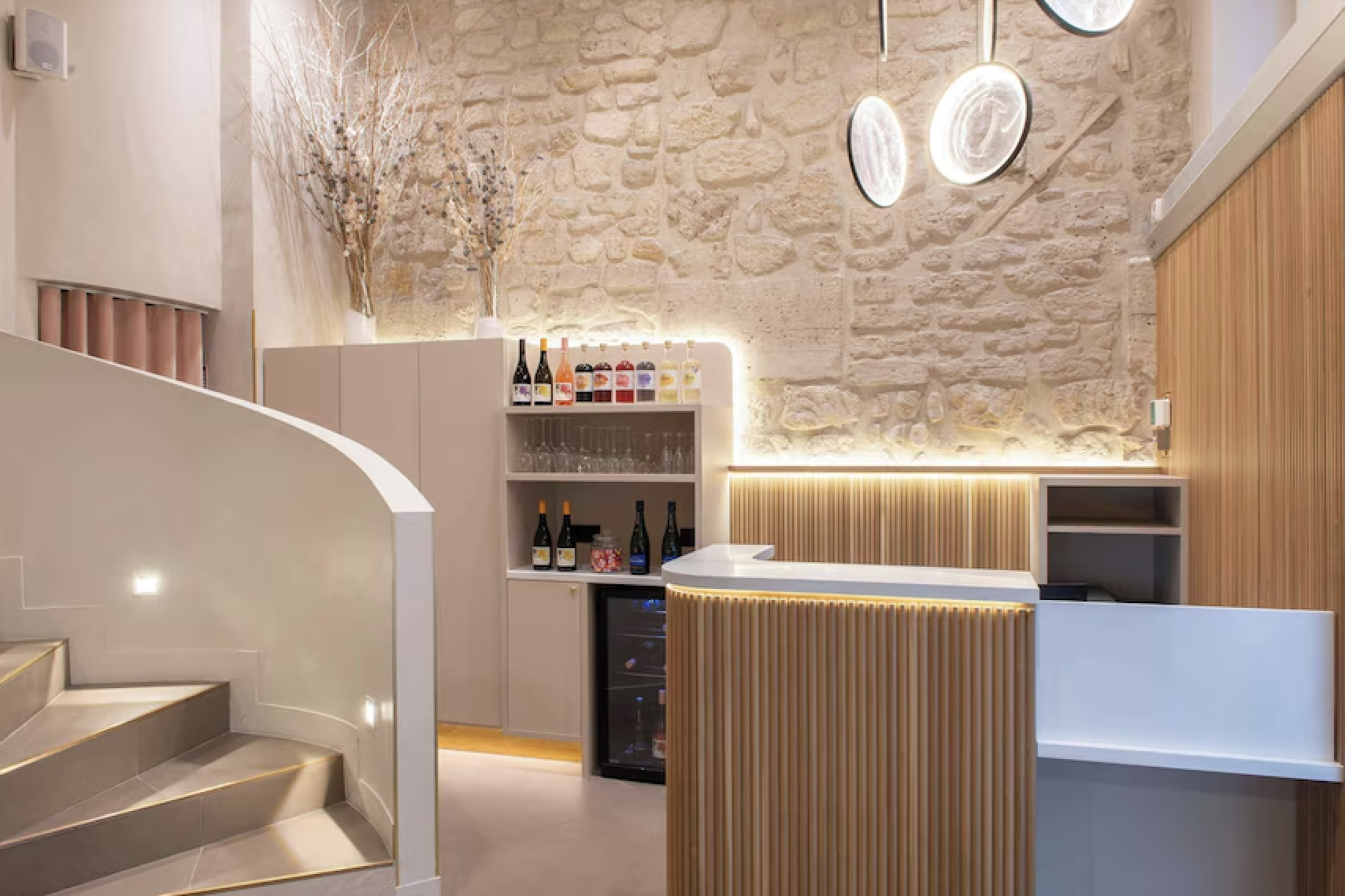 Modern bar area with a curved wooden counter, shelves with wine bottles and glasses, and decorative lighting on a textured stone wall.