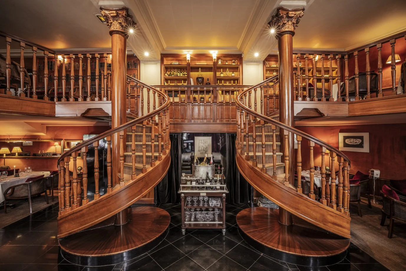 Elegant two-story restaurant with wooden staircase, railings, and columns, featuring a bar area with glassware and bottles, warm lighting, and dining tables with chairs.