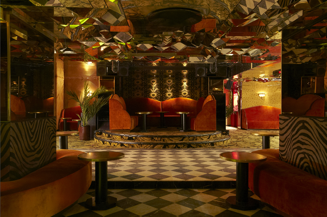 An interior view of a dimly lit lounge or nightclub with plush orange seating, a small elevated stage with matching seating, and reflective ceiling decor. The floor has a diamond-patterned black and white tile, and there are potted plants adding to the ambiance.
