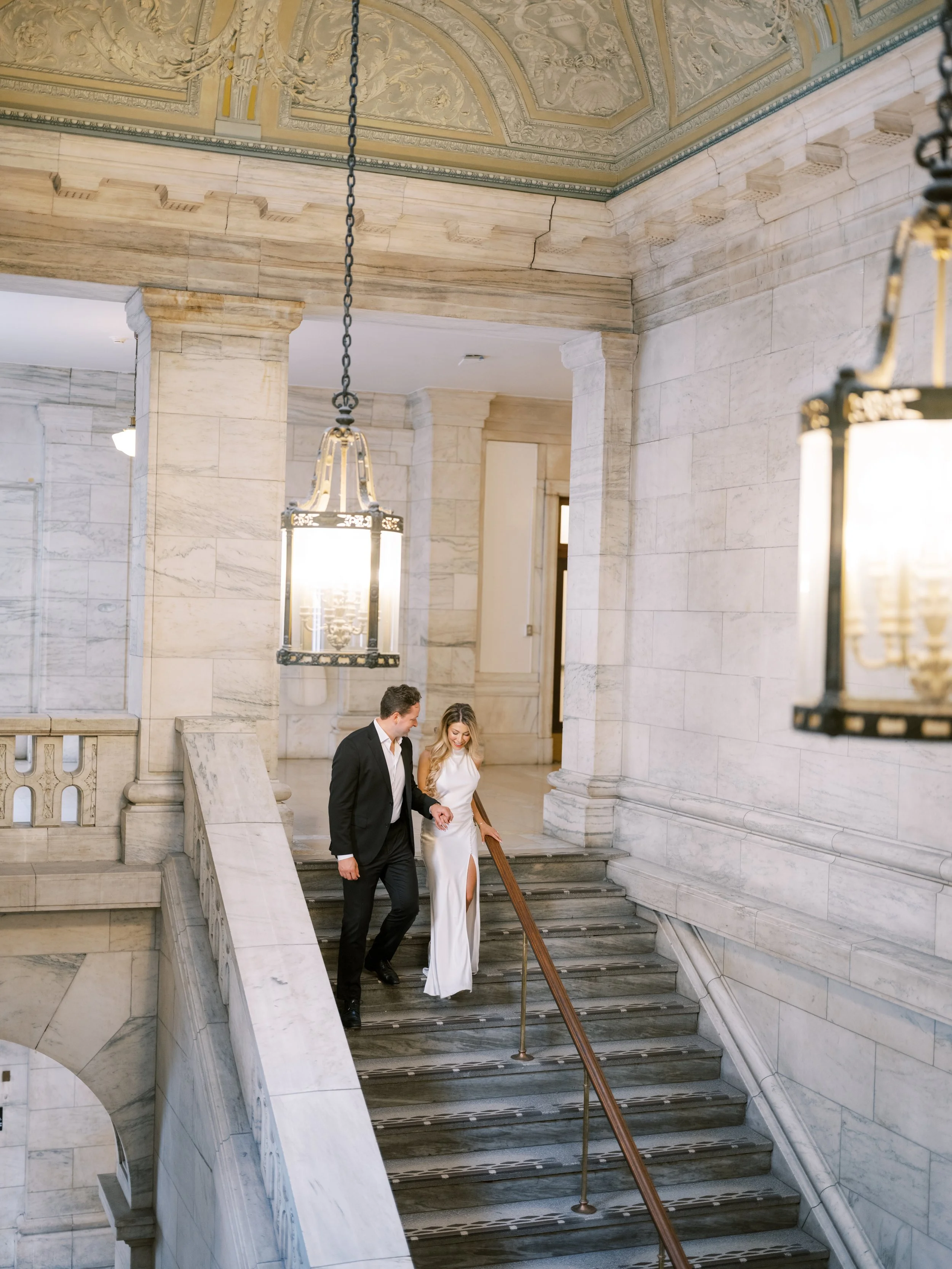 A couple, dressed in formal attire, walking down a marble staircase inside a grand, historic building with ornate ceilings and hanging chandeliers. New York Public Library. NYPL. Samantha Daniell & Michael Moore. 