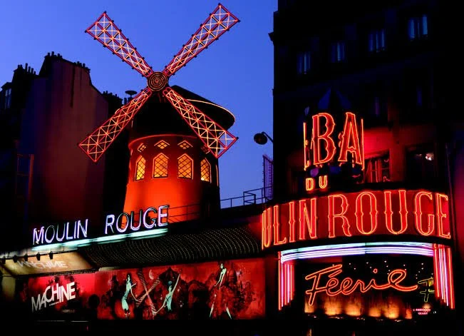 Night view of the Moulin Rouge in Paris, featuring a large, illuminated red windmill with spinning blades, neon signs including "Moulin Rouge" and "Féerie," and a lively atmosphere.
