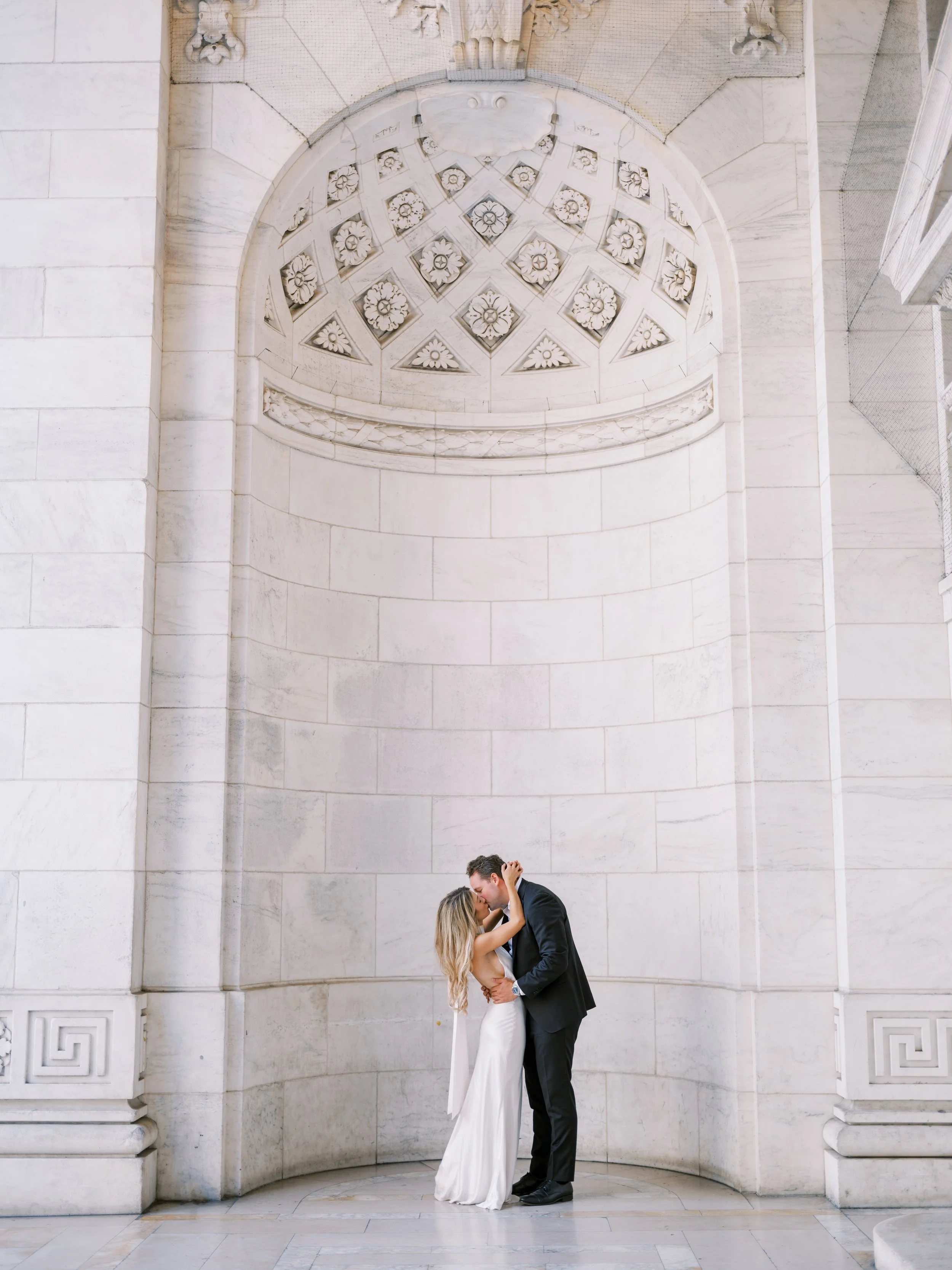 A bride and groom share an intimate kiss beneath a large, decorative white marble archway with intricate floral cutouts, in an elegant, classical architectural setting.