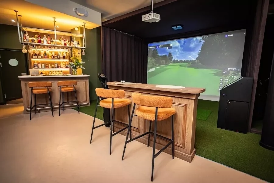 Indoor golf simulation room with a large screen showing a golf course, a wooden bar with four stools, and a small bar area with bottles and glasses.
