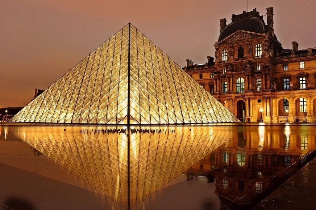 Illuminated glass pyramid sculpture at night with a historic building in the background and its reflection in the water