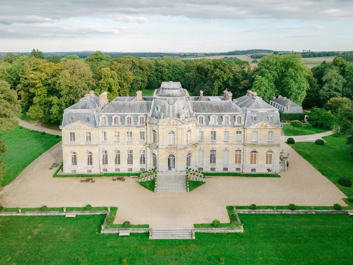 A large, historic mansion with elaborate architecture, surrounded by lush green lawns, trees, and gravel walkways.
