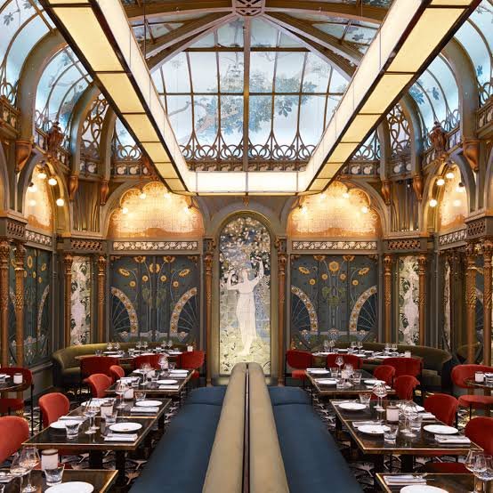 Elegant upscale restaurant with ornate decor, large glass ceiling, and classic artwork on walls, set up for dining with tables, glasses, and chairs.